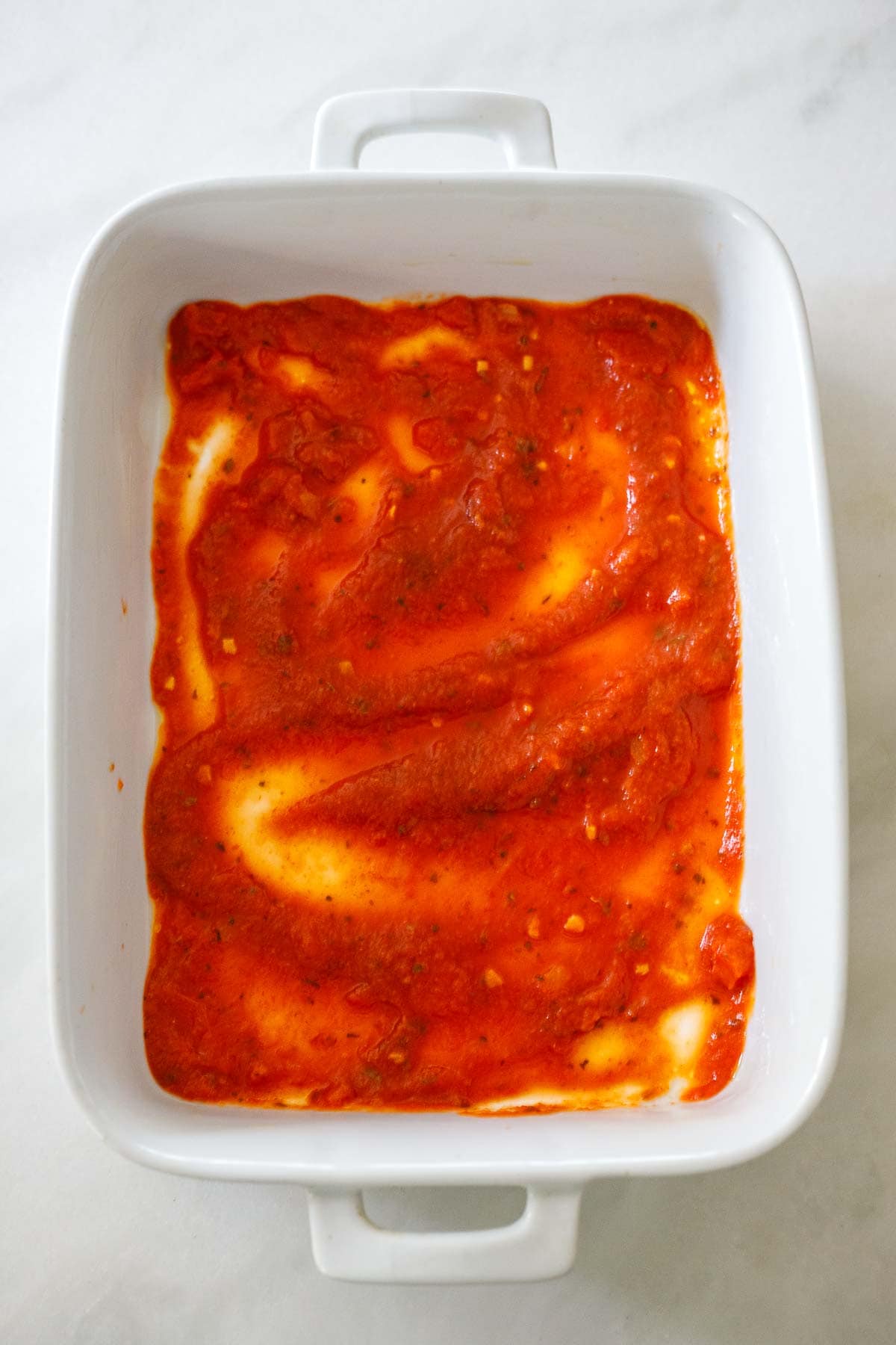 white casserole dish with layer of marinara sauce