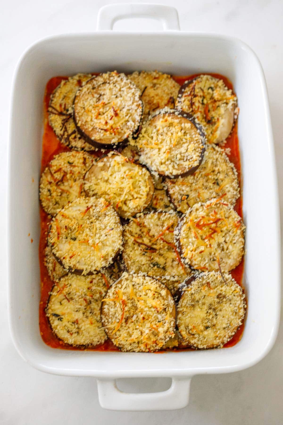 white casserole dish with baked eggplant on top of layer of marinara sauce 