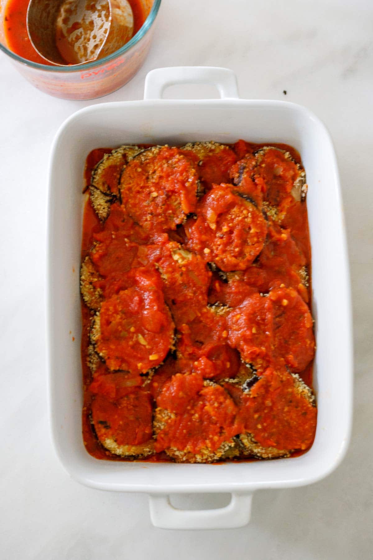 assembling eggplant parmesan with layer of marinara sauce over baked eggplant in white casserole dish
