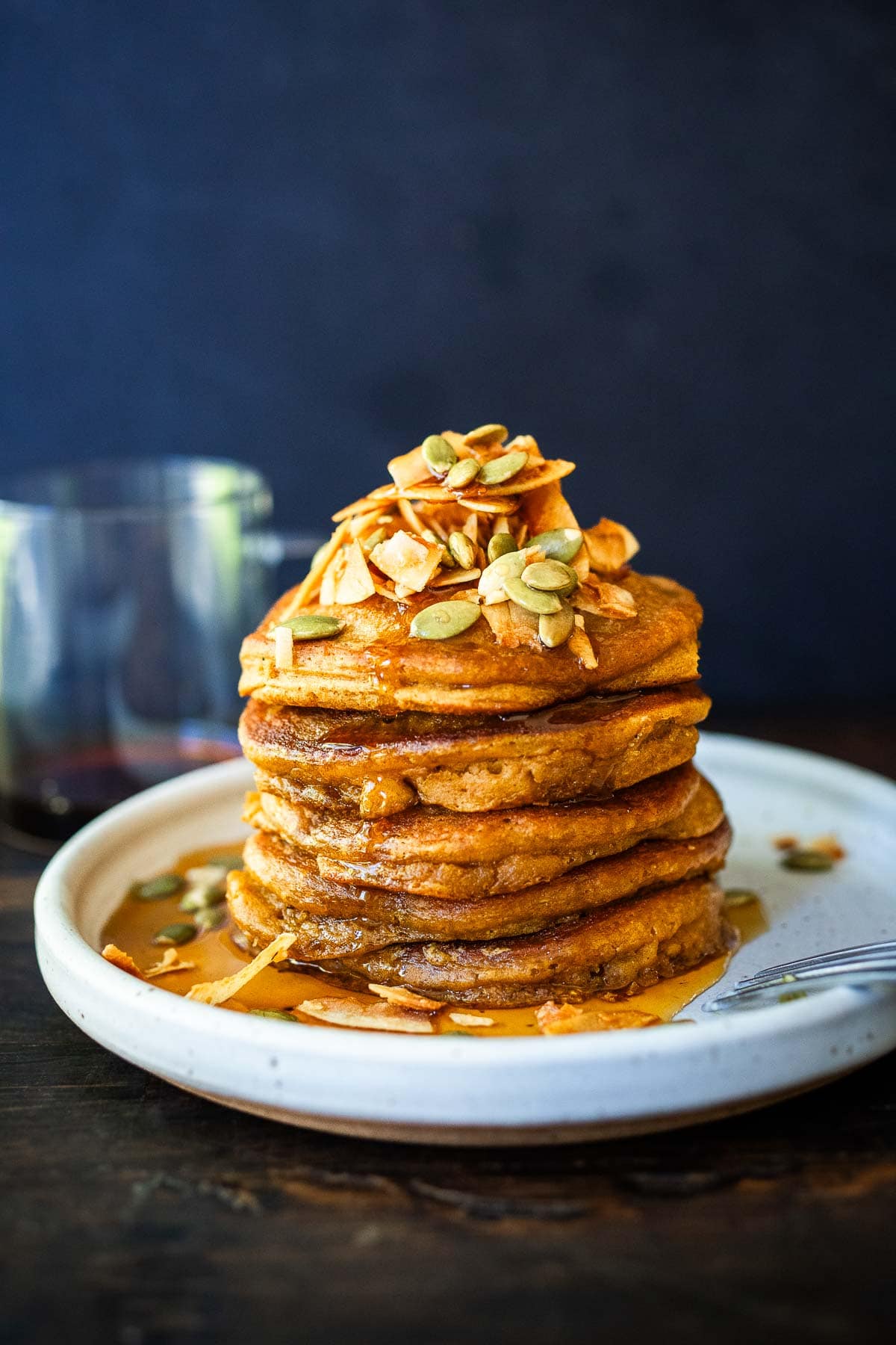 stack of fluffy pumpkin pancakes on plate with toasted coconut, pumpkin seeds, and maple syrup