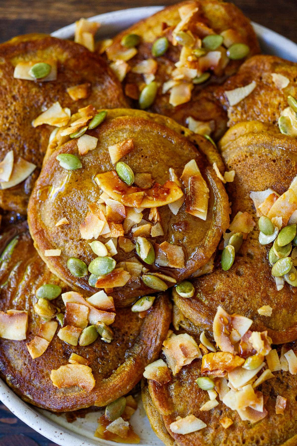 pile of fluffy pumpkin pancakes with toasted coconut, pumpkin seeds, and maple syrup
