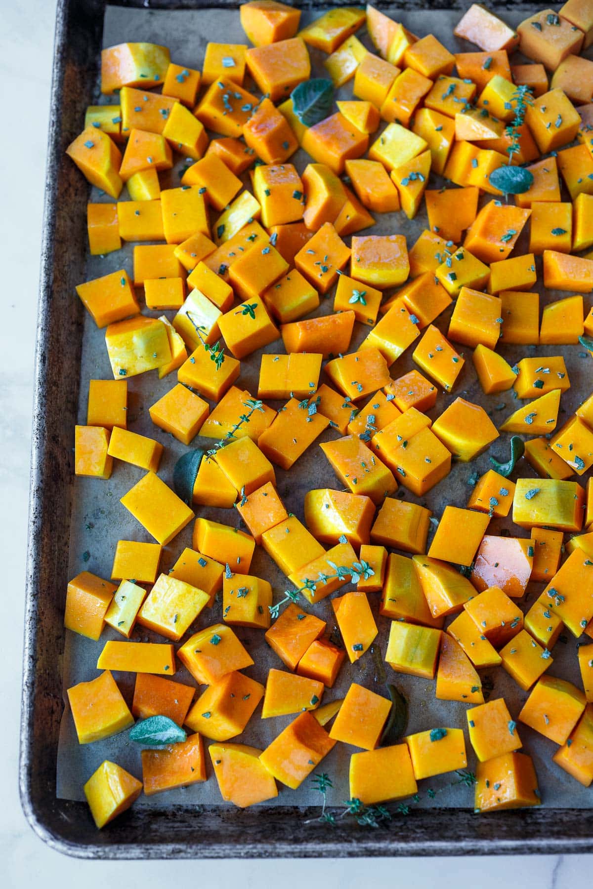 peeled and cubed butternut squash laid out on parchment lined baking sheet with fresh herbs