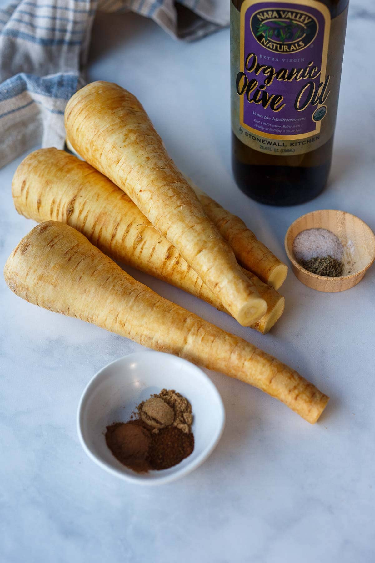 parsnips piled on table next to small dishes of spices and of salt and pepper with bottle of olive oil