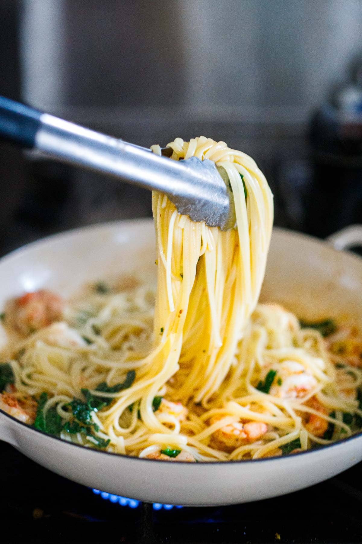 linguini pasta lifted by tongs in pan with shrimp and kale