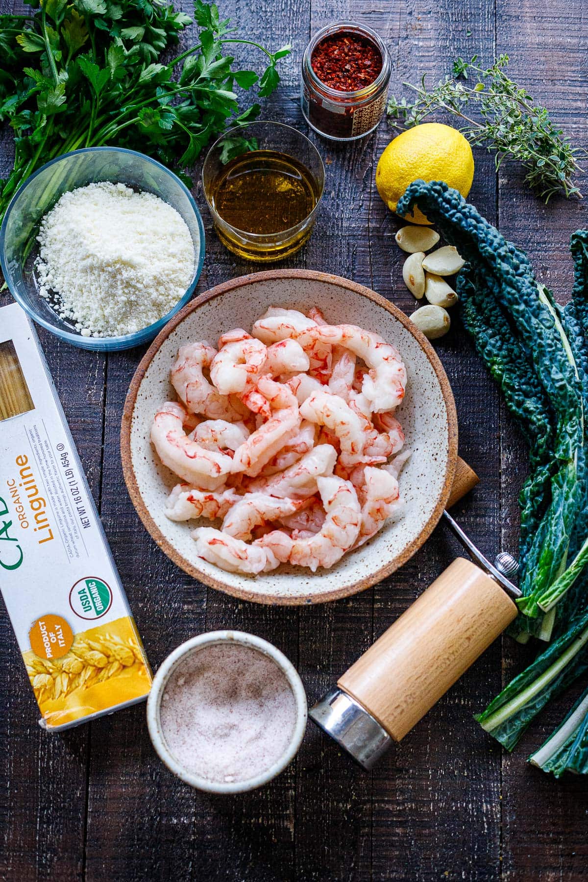 ingredients for shrimp pasta on wood table - bowl of shrimp, lacinato kale, garlic cloves, oil, lemon pecorino, aleppo, salt, linguine