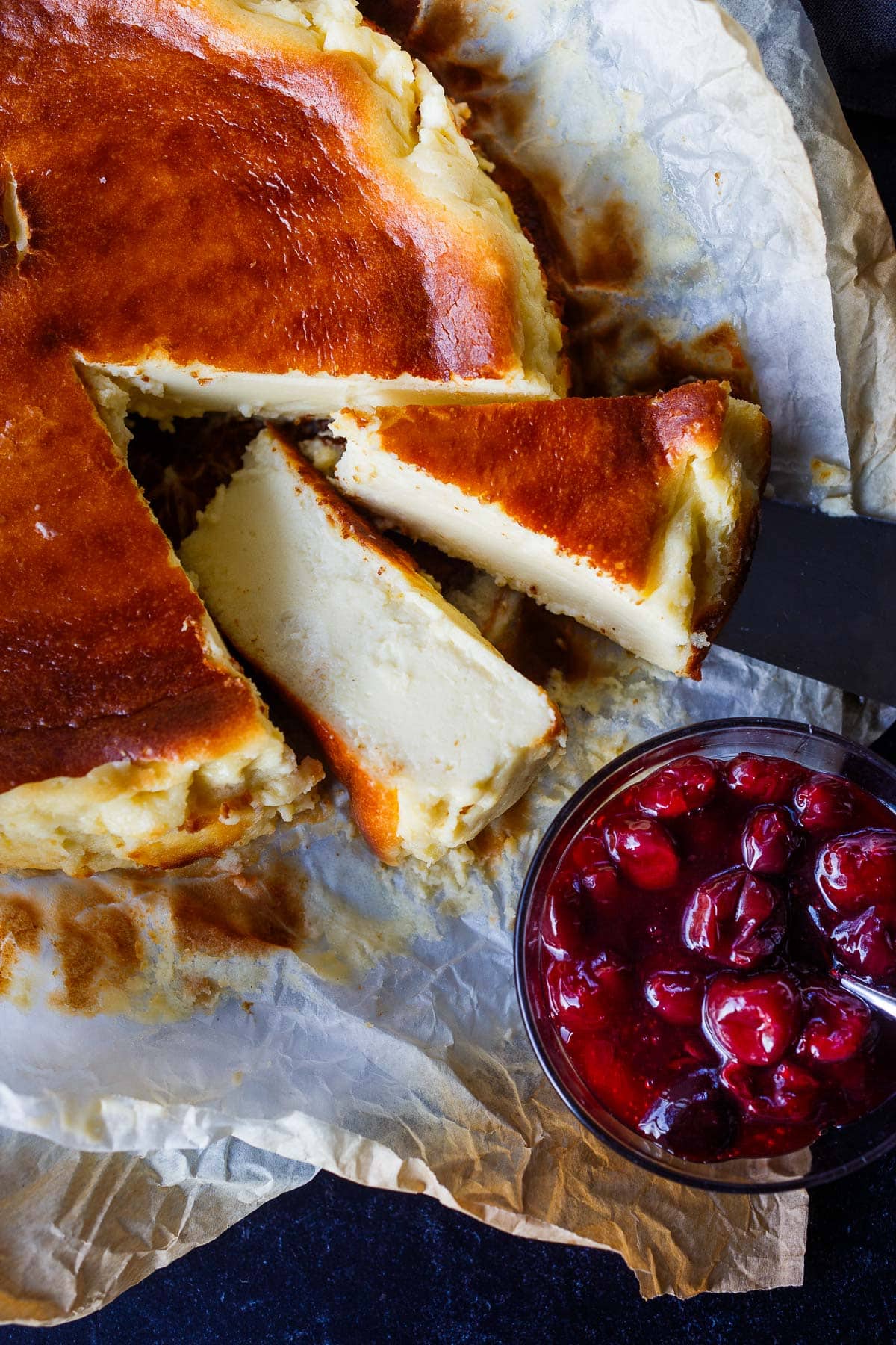 slices of burnt basque cheesecake cut out from round cheesecake on parchment paper next to berry compote