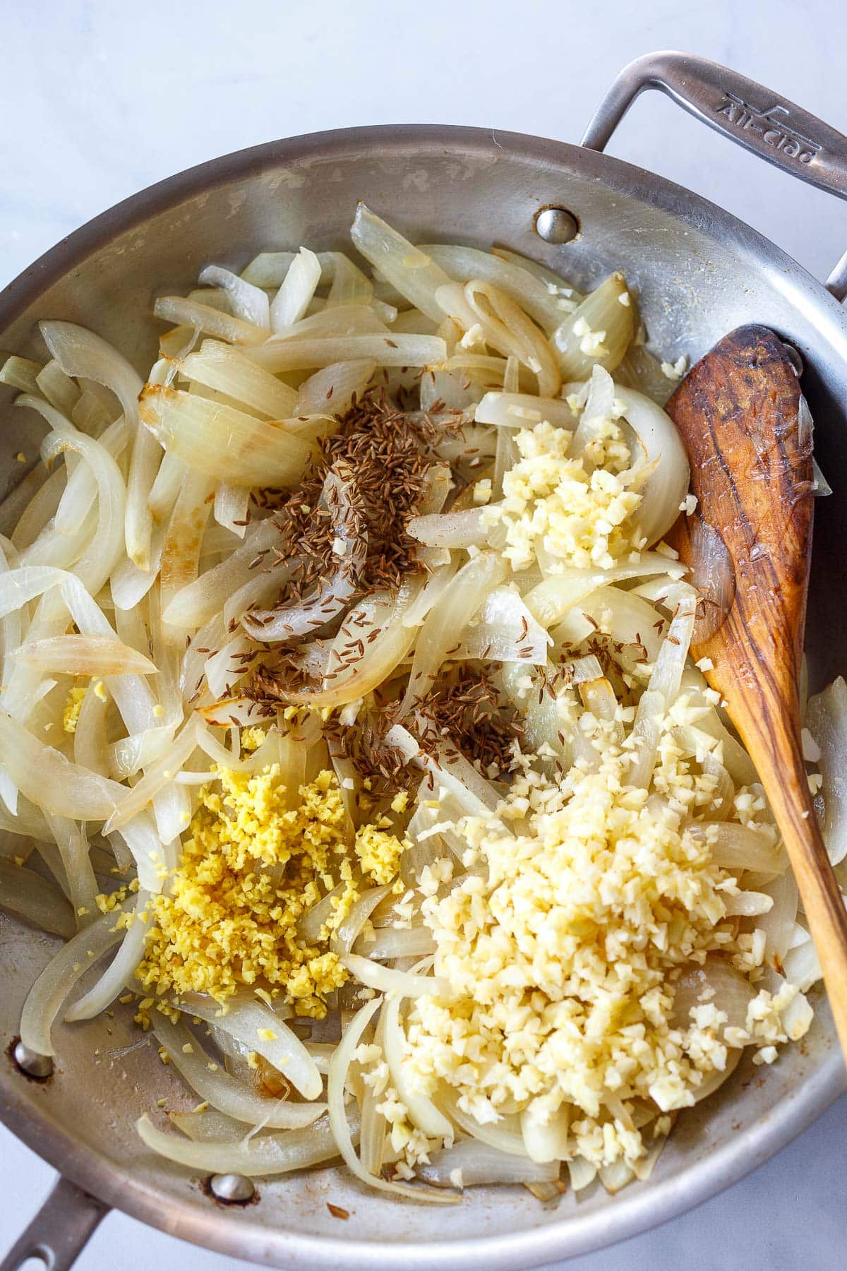 sliced onions in skillet with cumin seeds, garlic, and ginger tossed into pan with wooden spoon