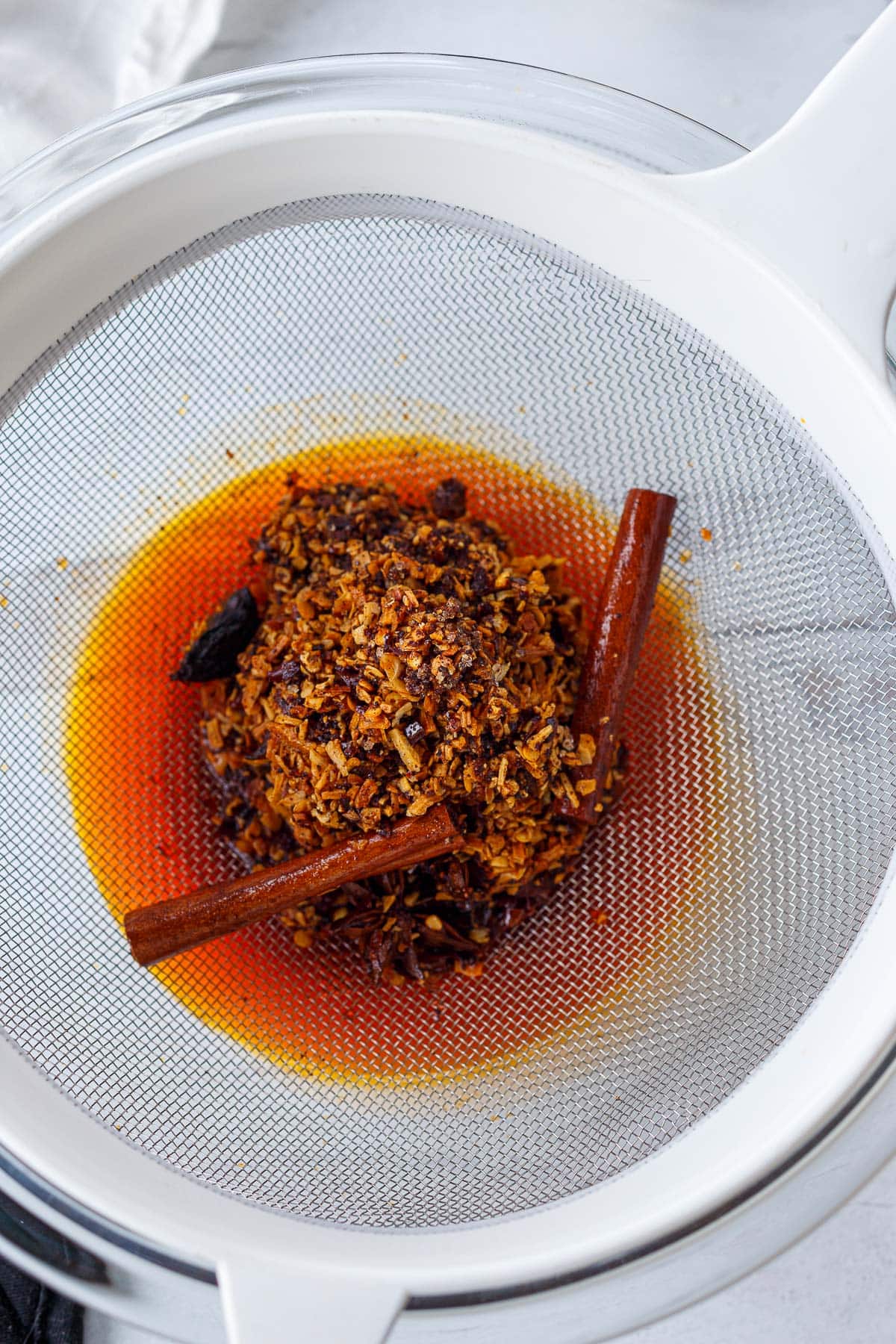 straining chili crisp through fine mesh sieve with cinnamon sticks, chilies, and aromatics