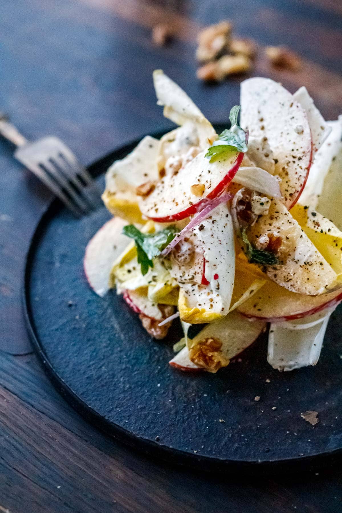 endive salad stacked on plate with apples and red onion, garnished with walnuts, parsley, black pepper.