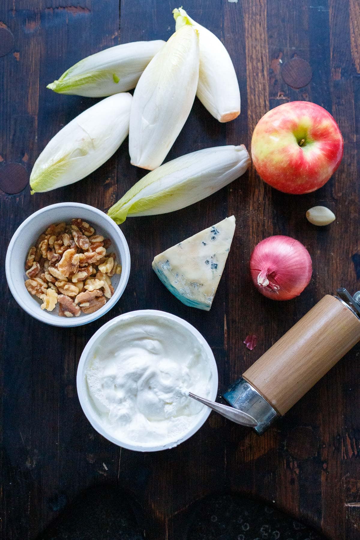 ingredients for endive salad on wood table - endive lettuce heads, apple, red onion, blue cheese, garlic, pepper shaker, walnuts, yogurt.