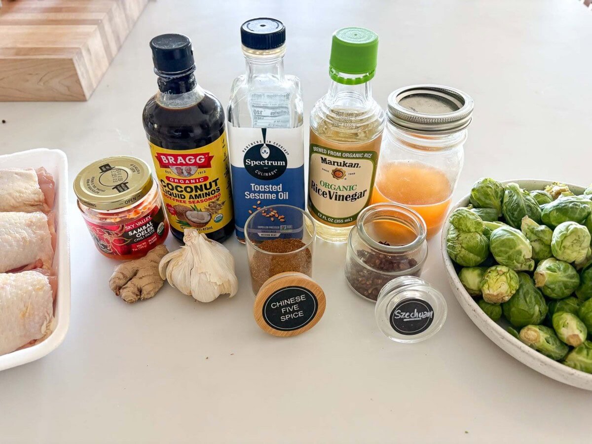 ingredients for sheet pan chicken - sambal oelek, ginger, garlic, coconut aminos, toasted sesame oil, Chinese five spice, rice vinegar, szechuan, and honey, next to bowl of brussels sprouts.
