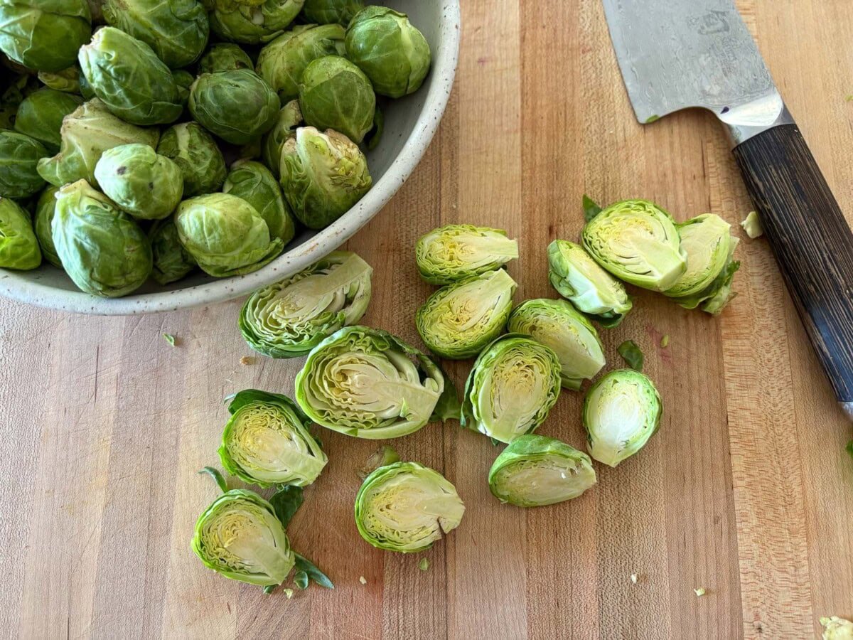 brussels sprouts halved on wood board with sharp knife and bowl of whole brussels sprouts.
