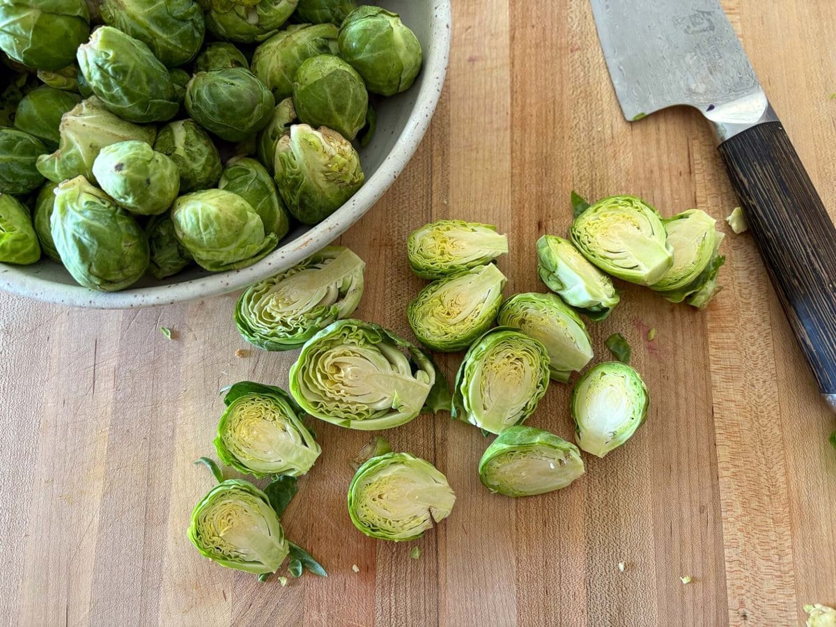 brussels sprouts halved on wood board with sharp knife and bowl of whole brussels sprouts.