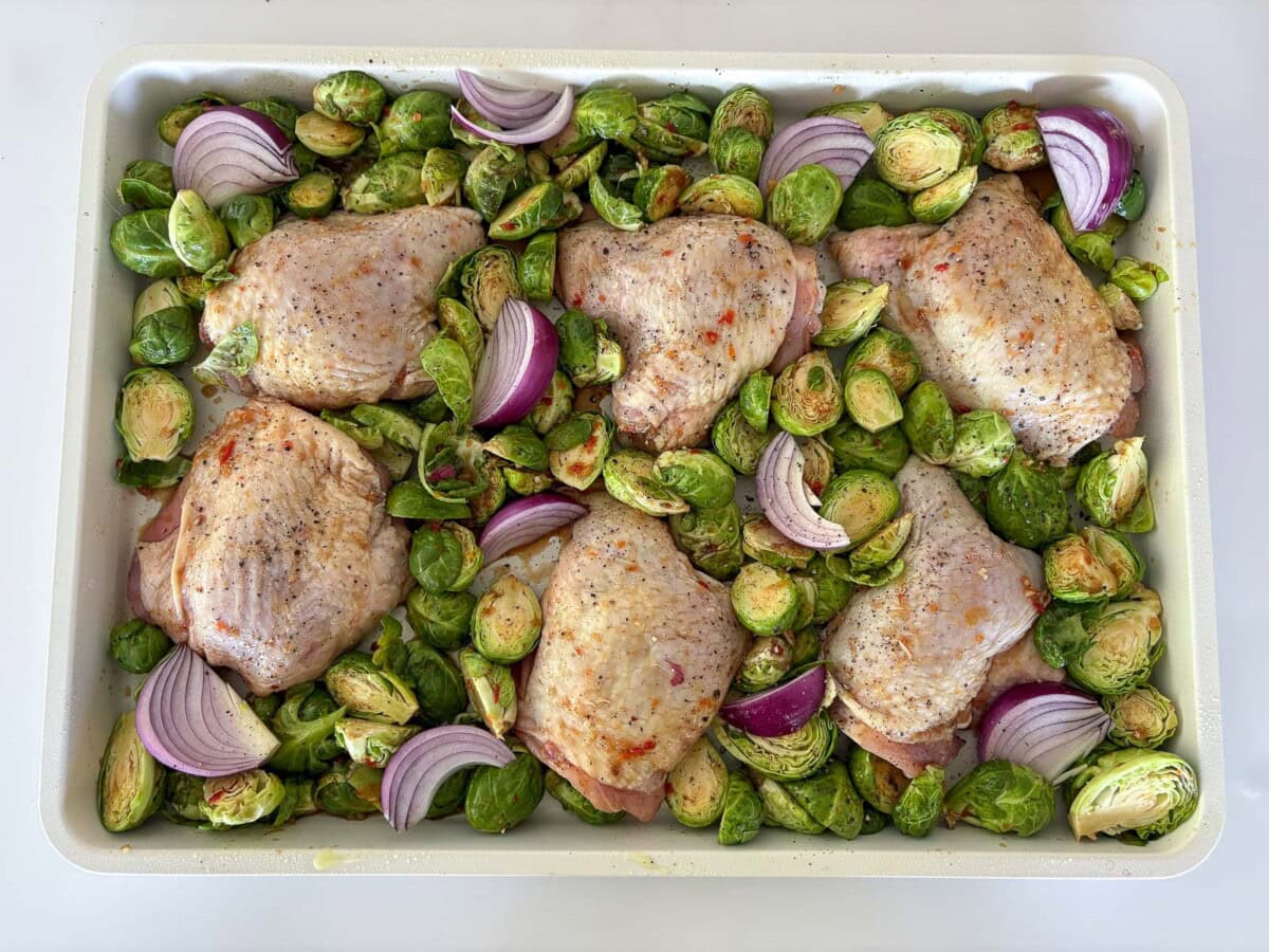 baking sheet with marinated chicken thighs, red onion wedges, and halved brussels sprouts.