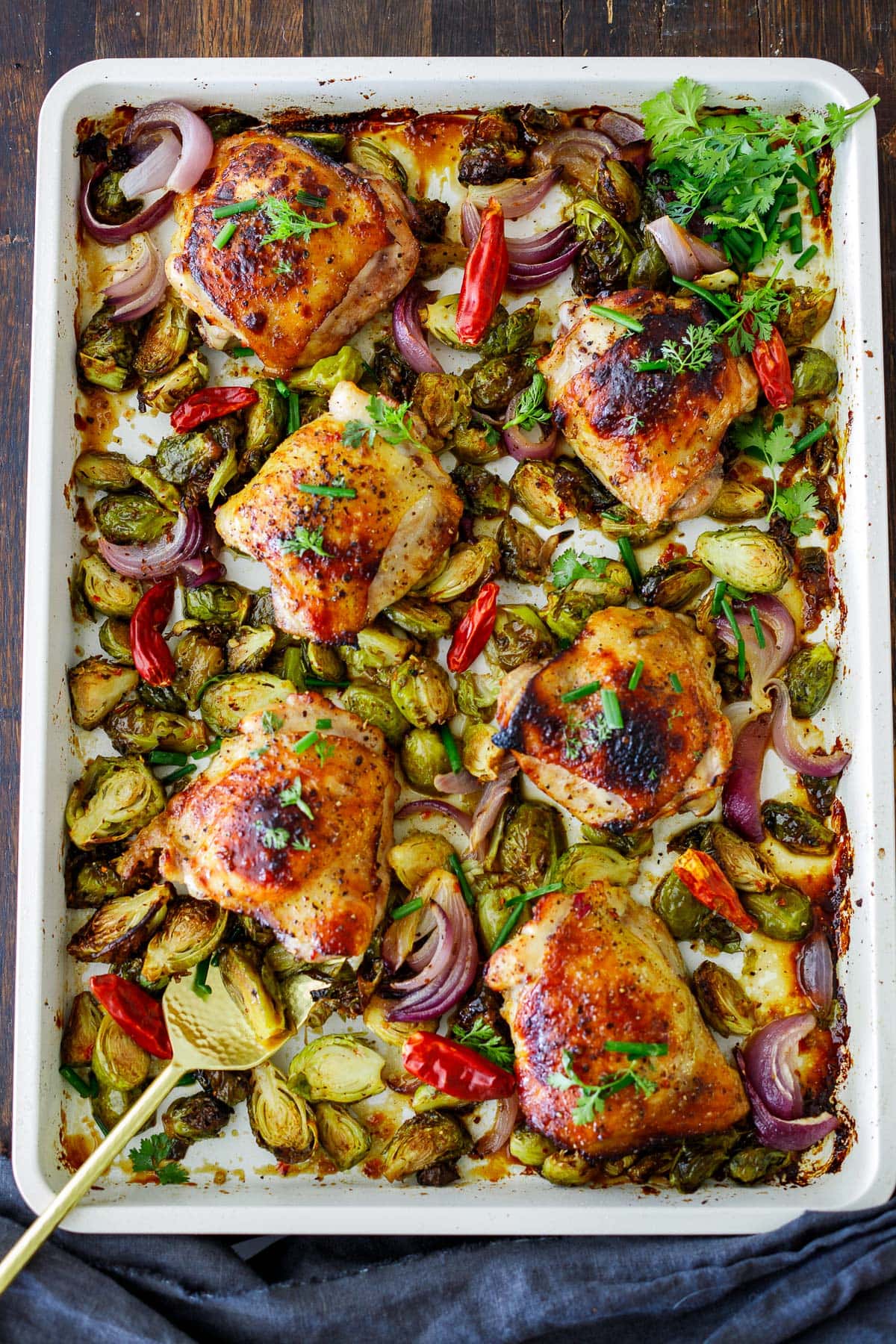 white baking sheet with baked szechuan chicken with roasted brussels sprouts, red onions, and red chilies.