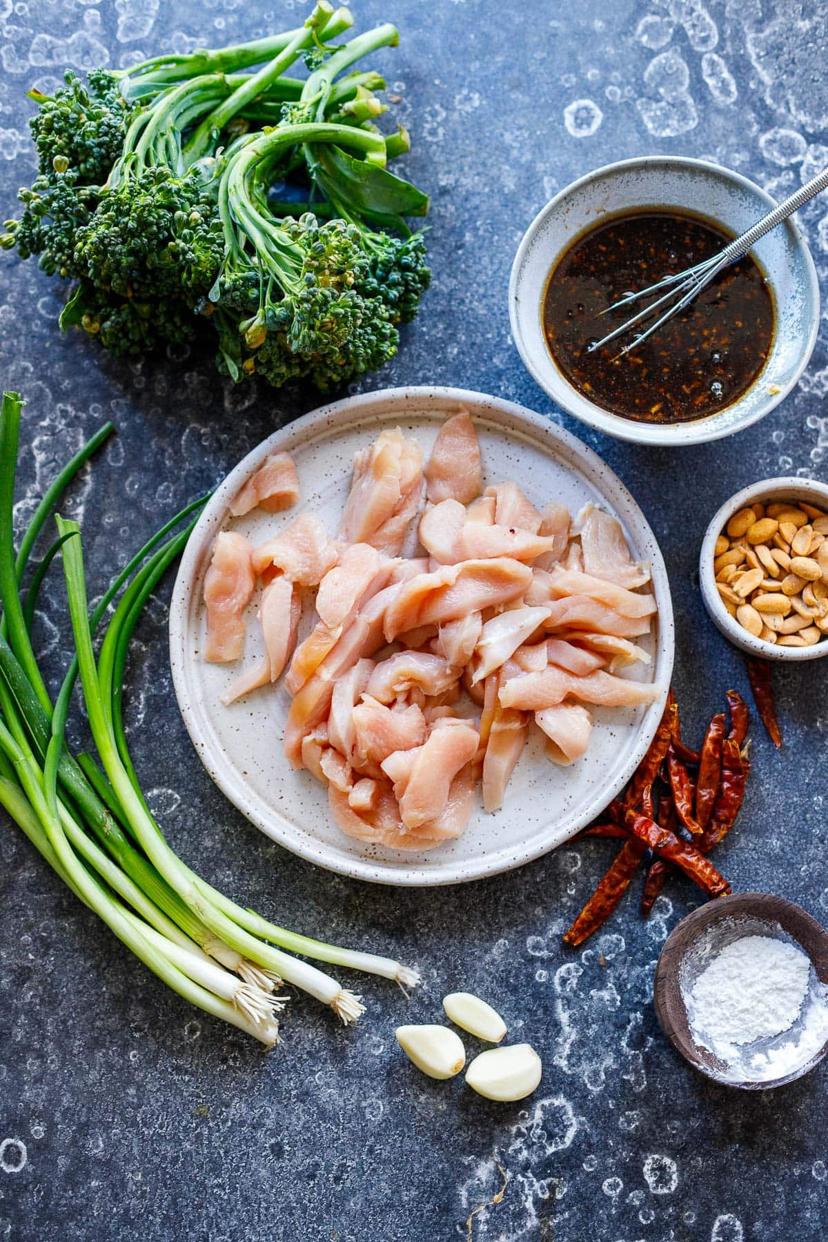 ingredients for szechuan chicken laid out - chicken sliced, scallions, garlic, peanuts, chilies, cornstarch, broccolini, szechuan sauce