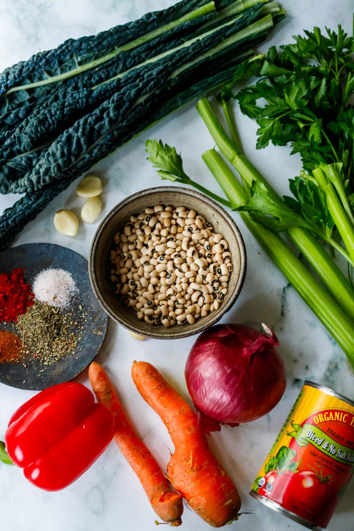 ingredients to make black eyed pea soup on marble surface - celery, carrots, onion, pepper, diced tomatoes, spices, kale, garlic.