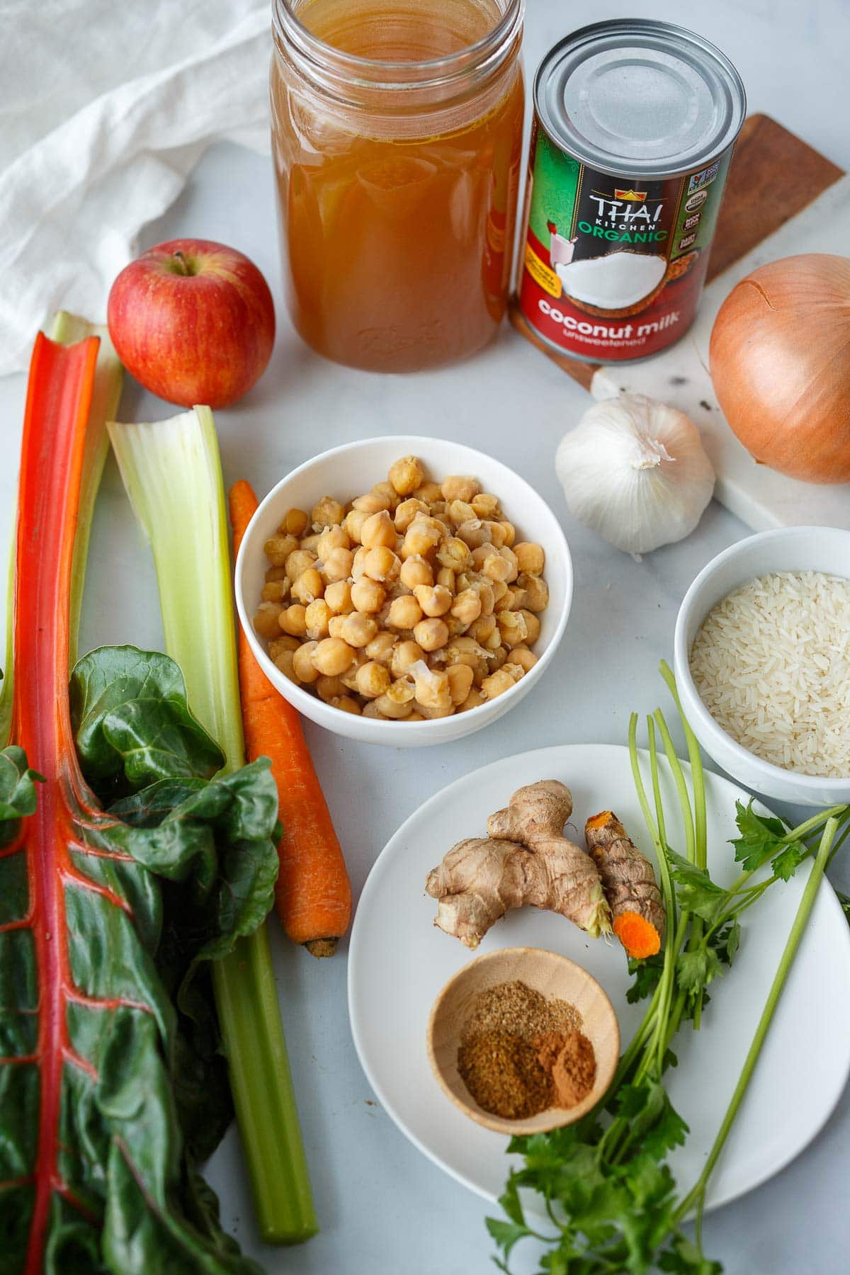ingredients for creamy chickpea soup laid out - chickpeas, coconut milk, onion, garlic, apple, broth, chard, celery, carrot, rice, turmeric, ginger, spices, parsley.