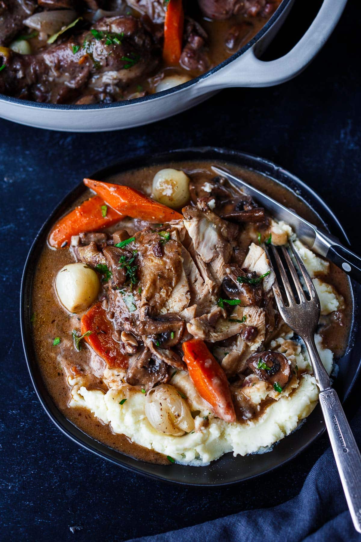 plate with coq au vin served over mashed potatoes with carrots, pearl onions, and mushrooms- fork cutting in to show tender meat.