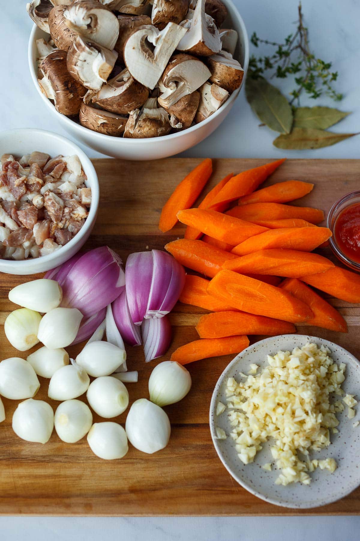 chopped carrots, sliced shallots, pearl onions, minced garlic, and chopped bacon on wood cutting board, next to chopped mushrooms, bay leaves, thyme.