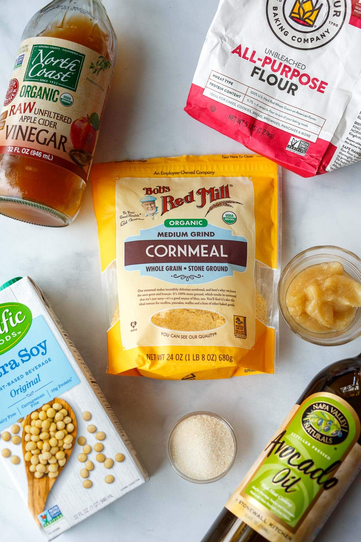 ingredients for vegan cornbread muffins laid out on table - cornmeal, flour, applesauce, vinegar, soy milk, sugar, avocado oil.