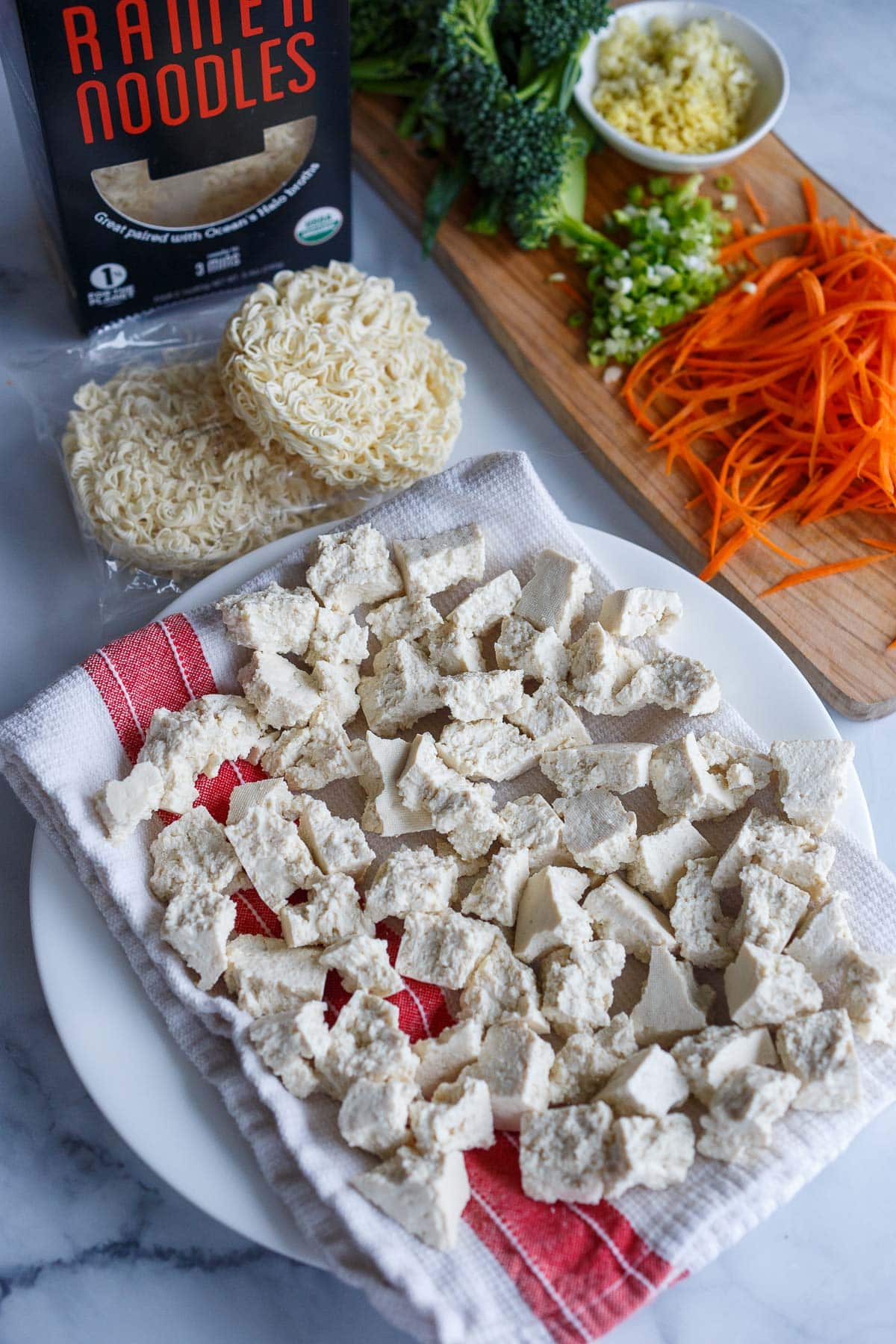 torn pieces of tofu laid out on plate with dish cloth beside other peanut butter ramen ingredients- noodles, veggies, garlic, ginger.