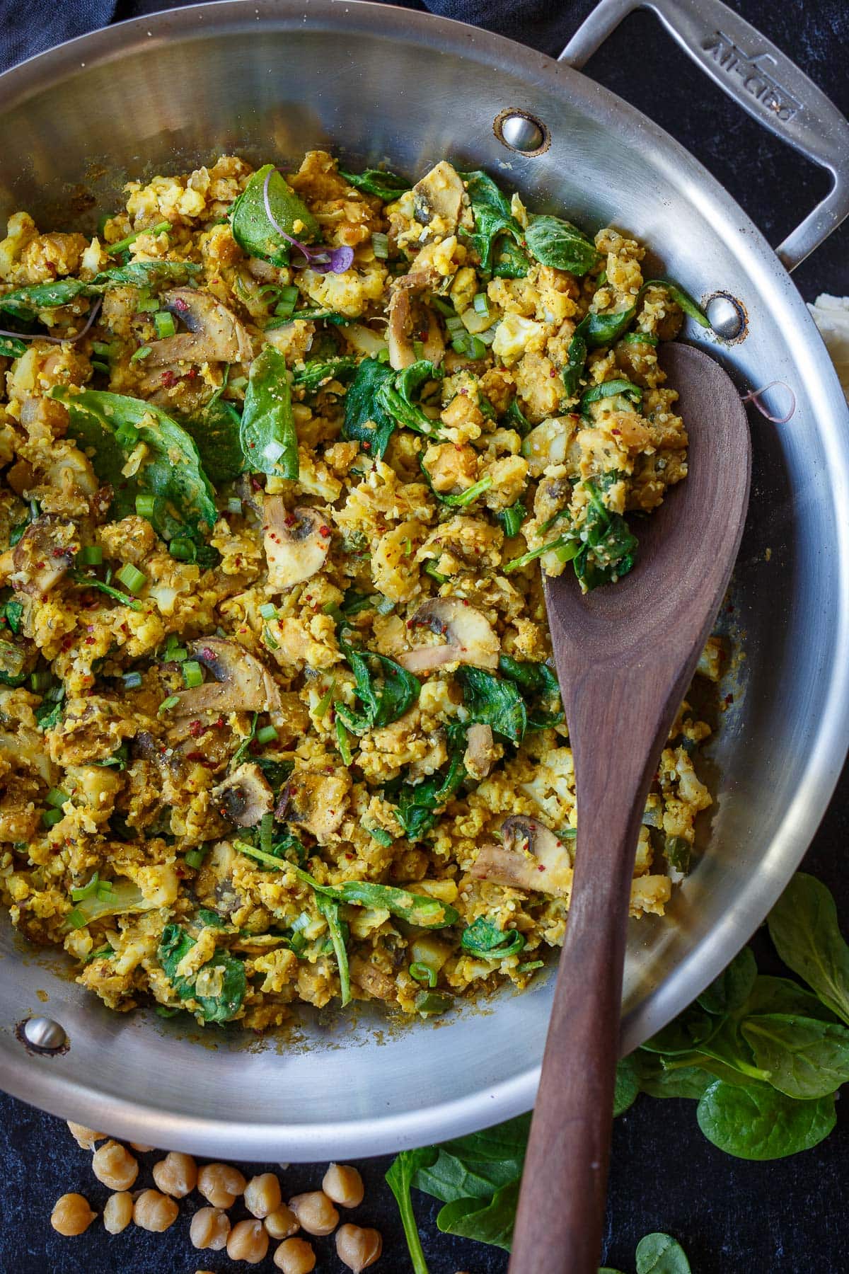 chickpea scramble in frying pan with spinach and mushrooms mixed in, stirred with wooden spoon.