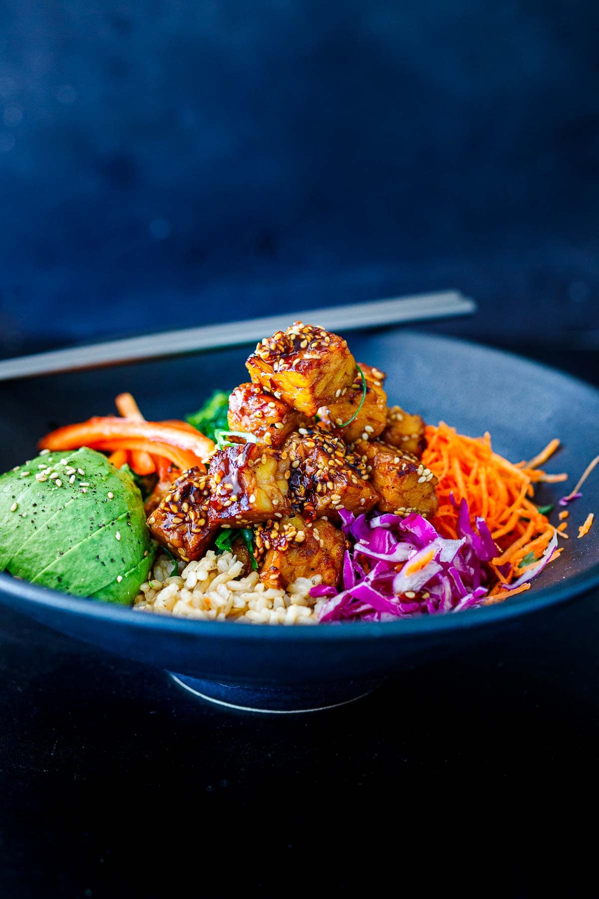 rice bowl with raw veggies, sliced avocado, and crispy sesame tempeh piled on top, garnished with sesame seeds.