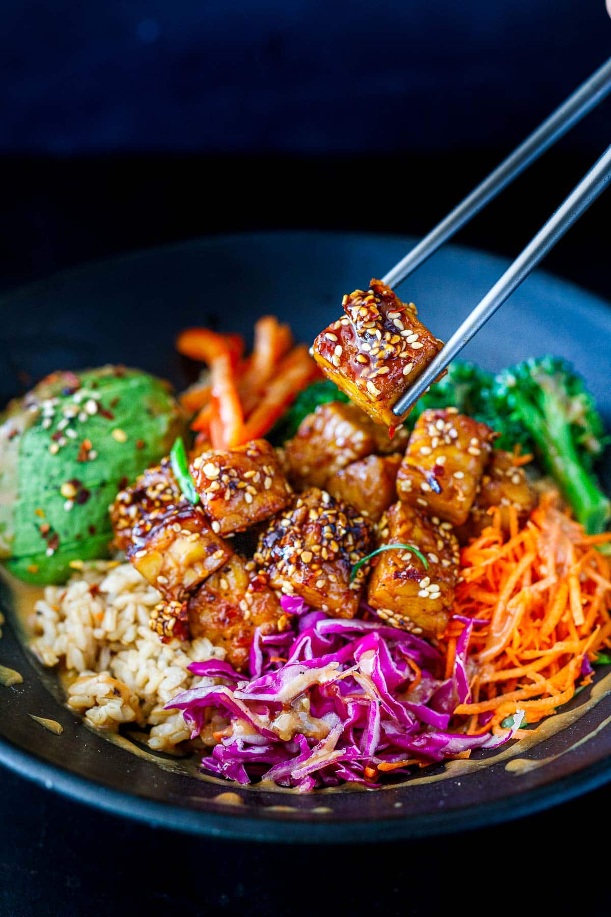 chopsticks holding up a piece of crispy sesame tempeh over a rice bowl with tempeh, raw veggies, rice, and avocado, garnished with sesame seeds.