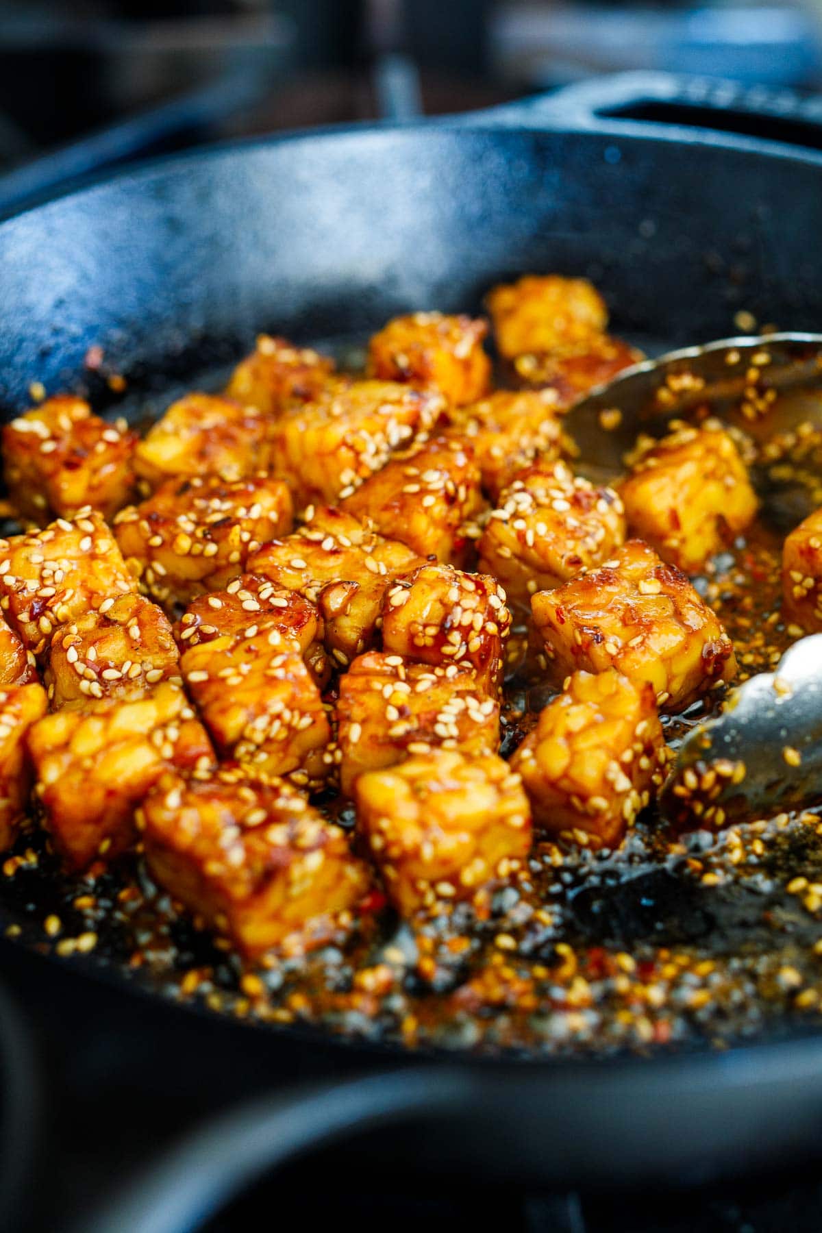 cubed tempeh frying in sticky sesame stir fry sauce in skillet with tongs, topped with sesame seeds.