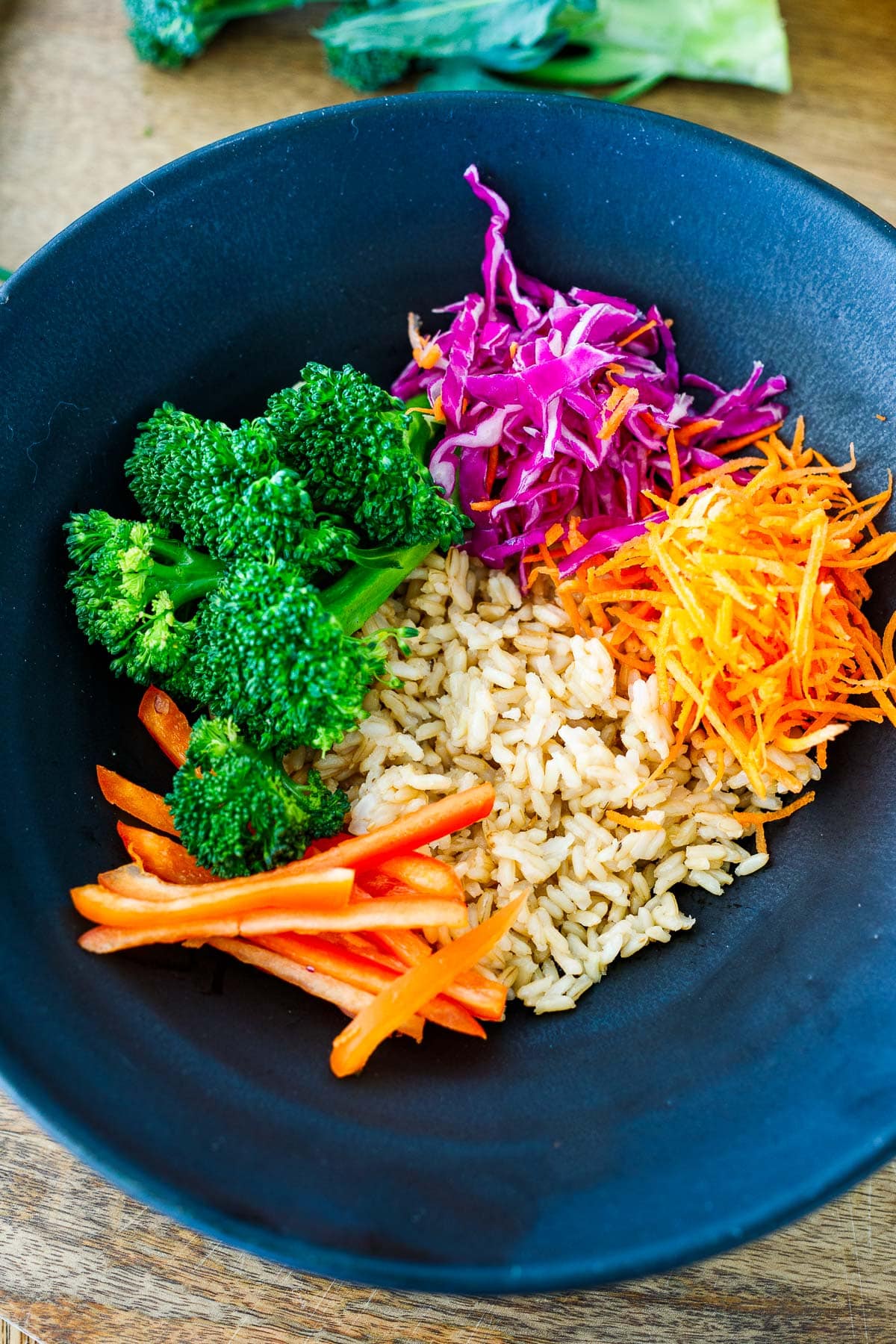 assembling rice bowl with brown rice, shredded carrots and cabbage, sliced bell pepper, and steamed broccoli.
