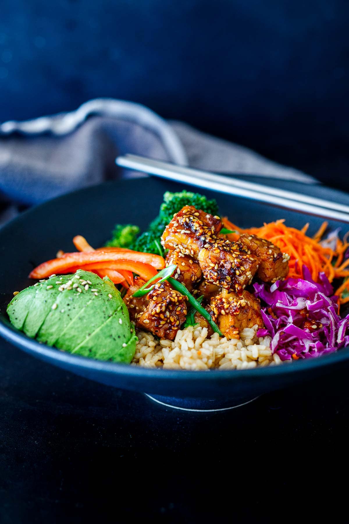 crispy sesame tempeh in bowl with rice and raw veggies, sliced avocado, topped with sesame seeds.