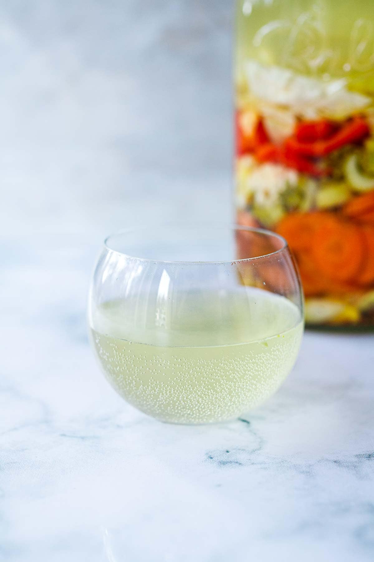 gut shot in a glass in front of jar of fermenting vegetables.