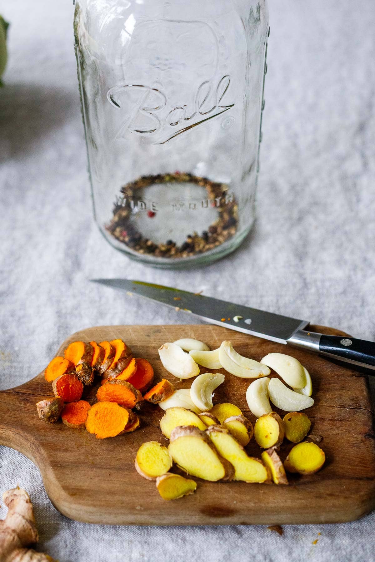 sliced ginger, turmeric, garlic cloves on wood cutting board next to glass jar with whole spices to make fermented gut shot.