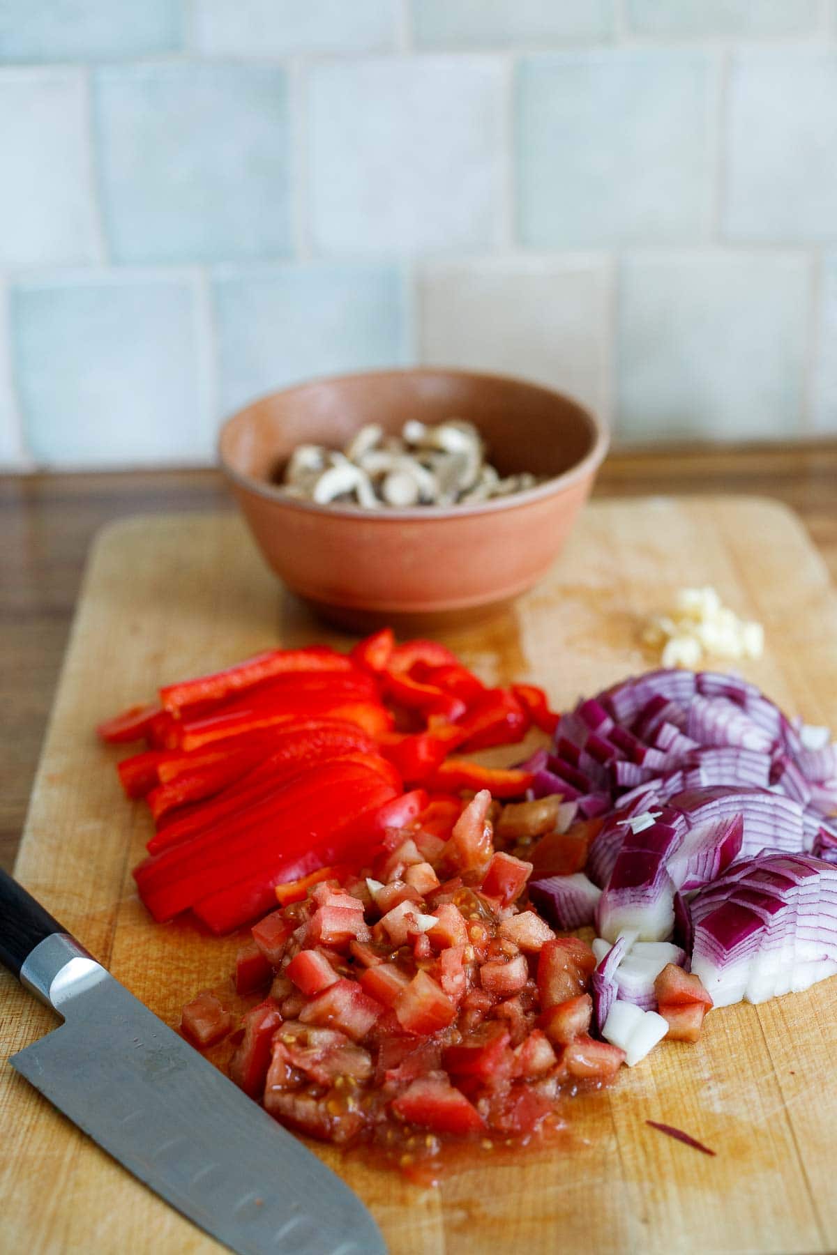 sliced red bell pepper, diced red onion, diced tomato and sliced mushrooms on wood cutting board with sharp knife.