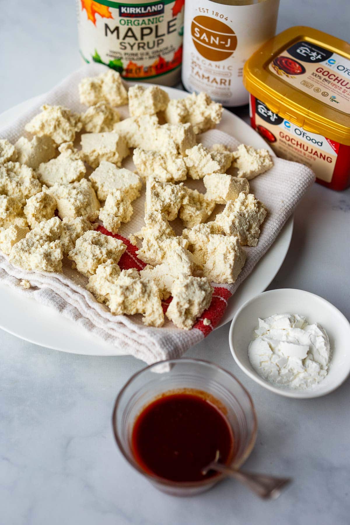 firm tofu broken into pieces on towel on plate, beside gochujang sauce and cornstarch. 