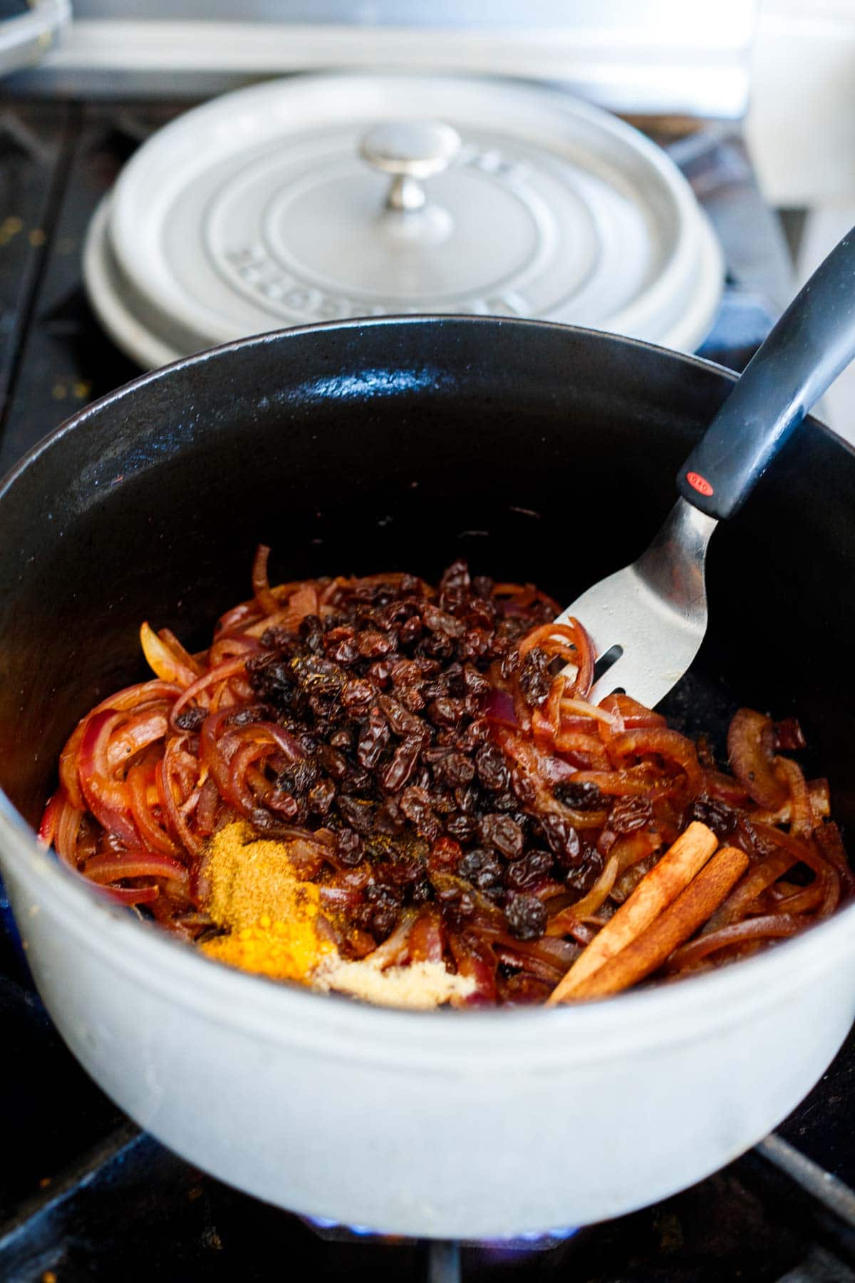 ingredients for Moroccan rice pilaf added to dutch oven with caramelized onions- raisins, spices, cinnamon sticks.