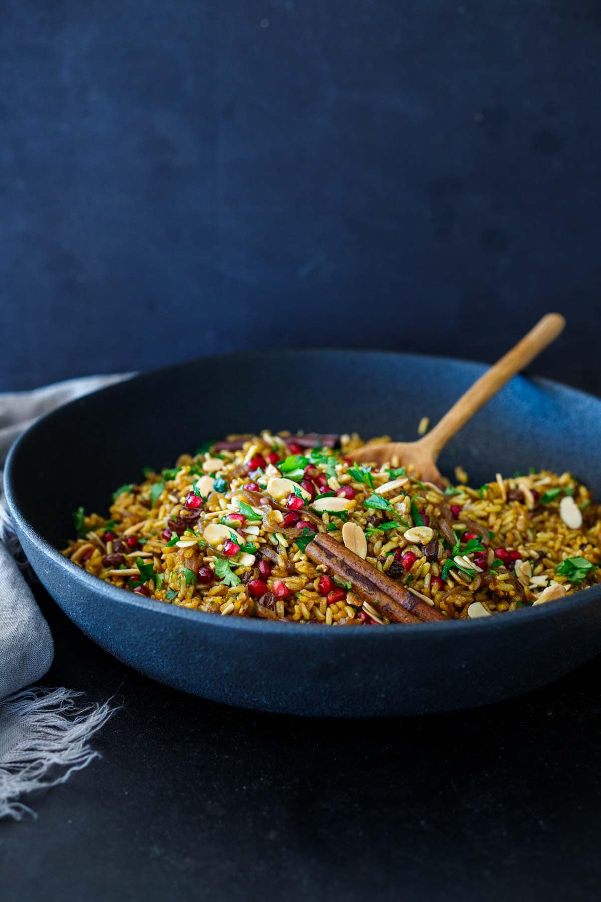 Moroccan rice pilaf in large serving bowl with wood spoon- served with cinnamon stick, minced parsley, slivered almonds, pomegranate seeds.