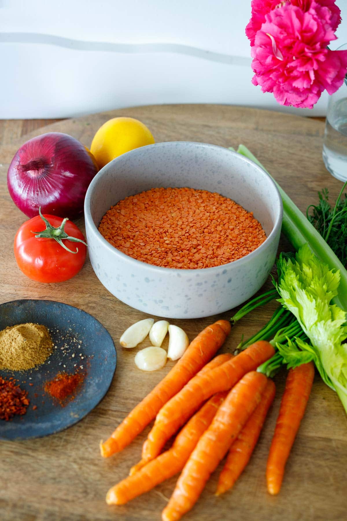 bowl of red lentils next to lemon, red onion, tomato, celery, carrots, garlic, and spices to make red lentil soup.