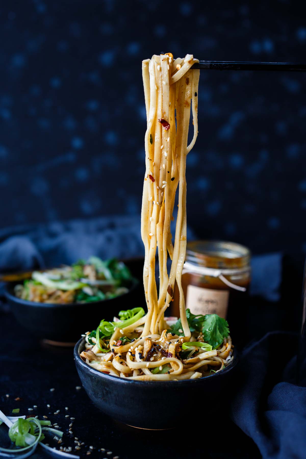 chopsticks holding up longevity noodles from bowl, which is garnished with cilantro, scallions, sesame seeds.