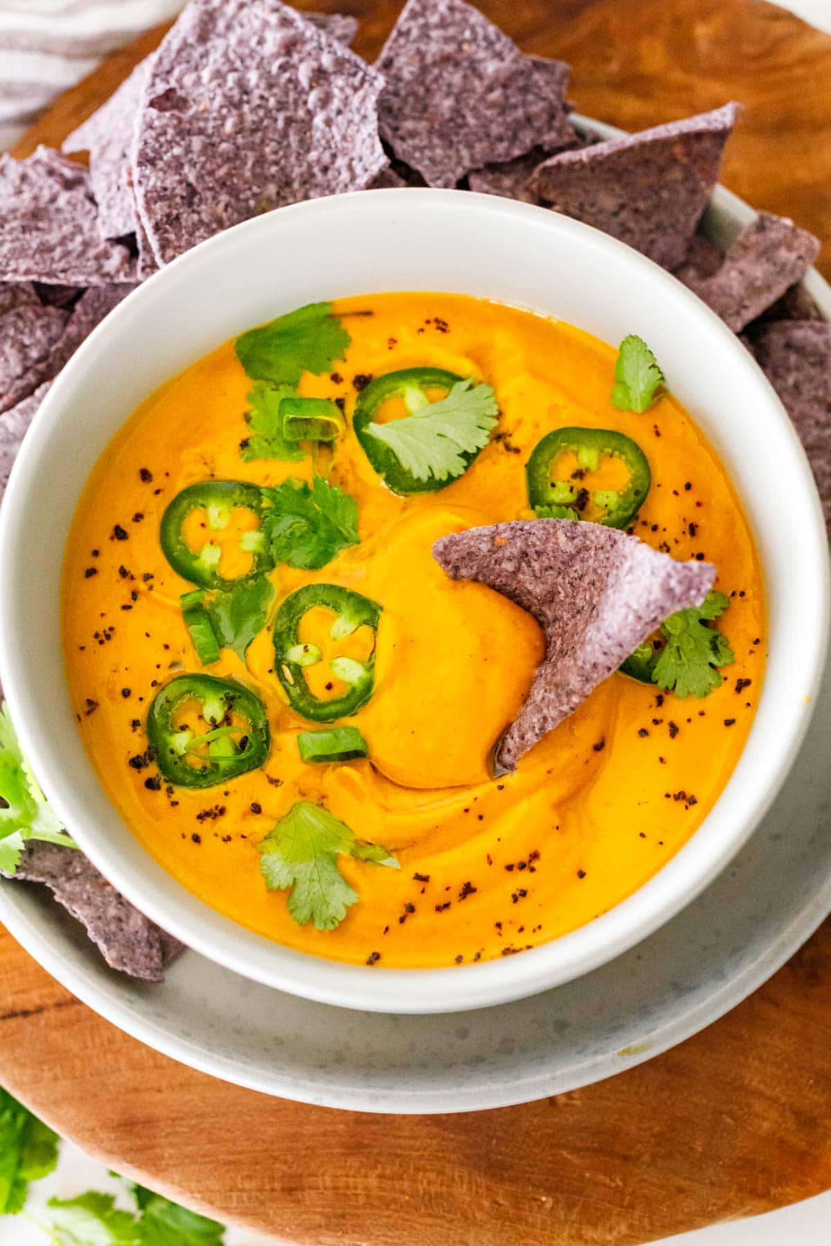 vegan queso in bowl with blue corn tortilla chip dipping, garnished with sliced jalapeños and scallions.