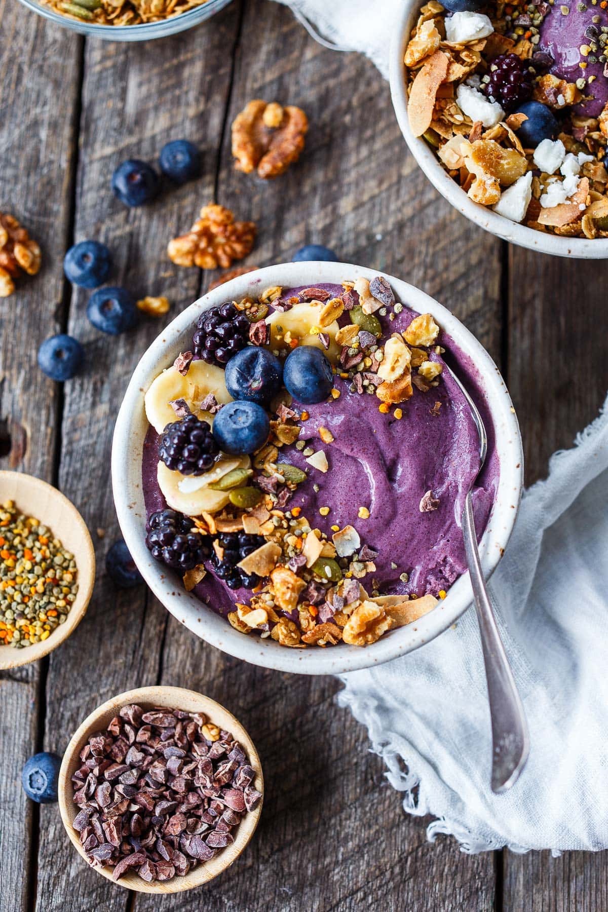 acai bowls with toppings- fresh blackberries, blueberries and sliced bananas, walnuts, granola, toasted coconut, beside bowl of cacao nibs and bee pollen.