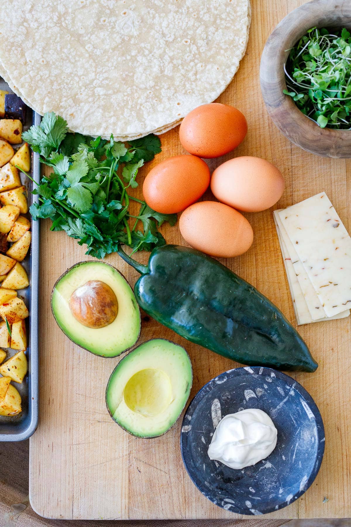 ingredients for vegetarian breakfast burritos - tortillas, microgreens, eggs, cheese, cilantro, poblano, avocado, potatoes, sour cream.