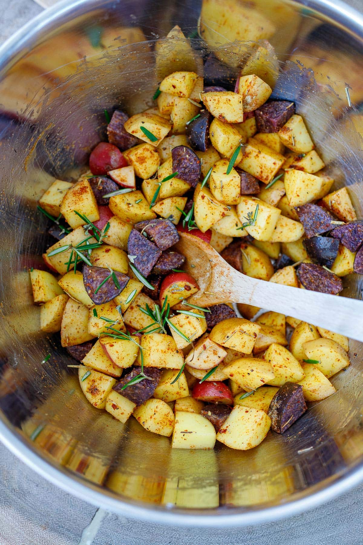 potatoes, spices, and herbs mixed together with wood spoon in large mixing bowl.