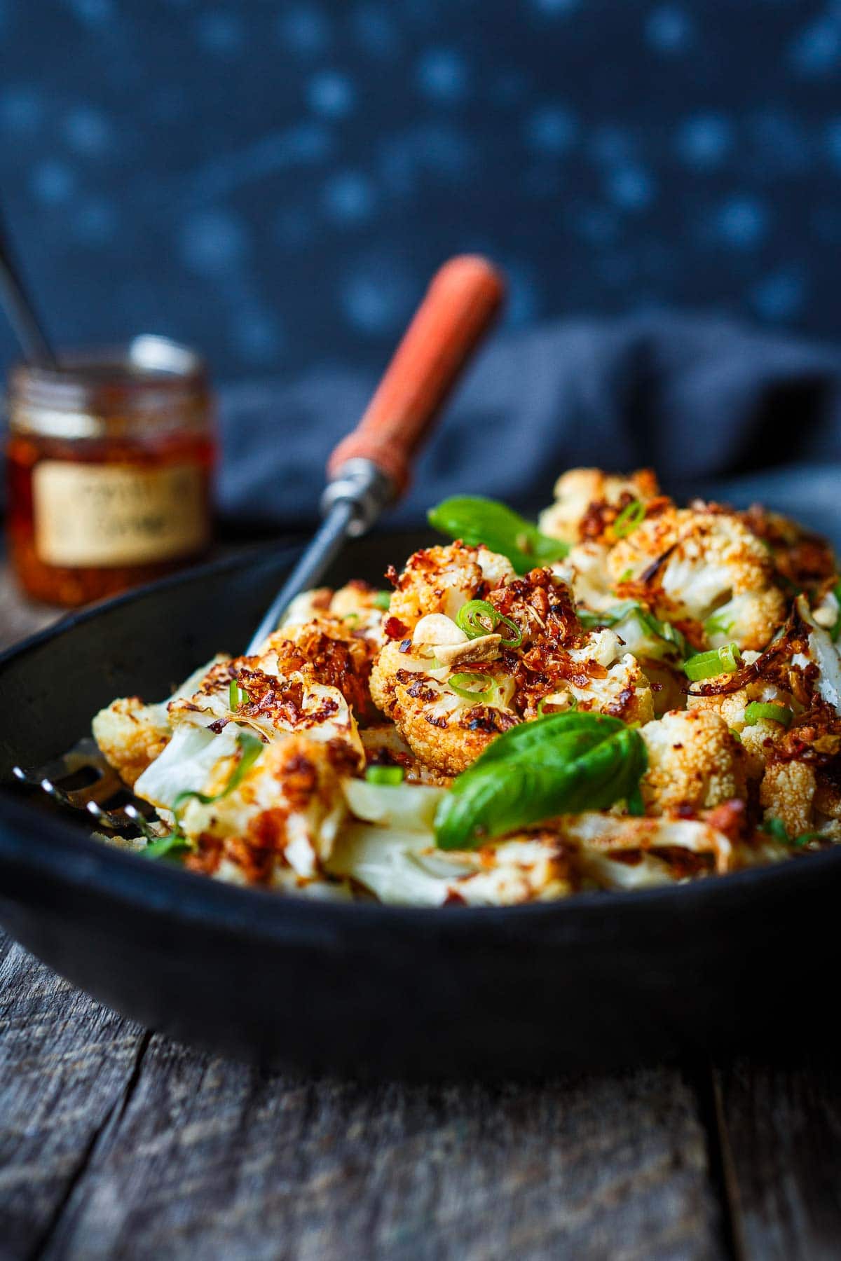 roasted chili crisp cauliflower in serving dish, garnished with basil leaves, scallions, crushed peanuts.