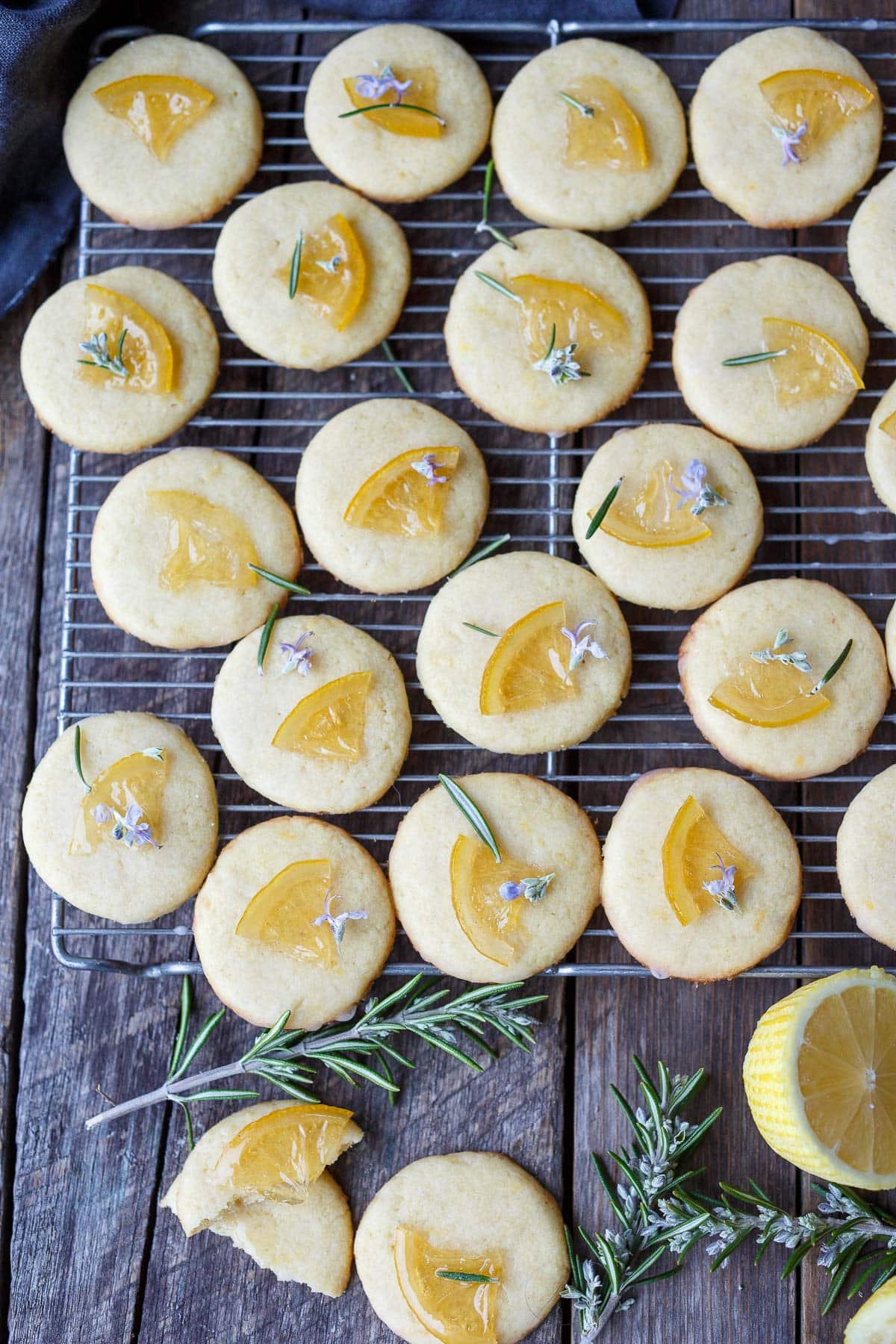 beautiful lemon cookies on wire rack, coated in lemon glaze and garnished with candied lemons, fresh rosemary and rosemary flowers.