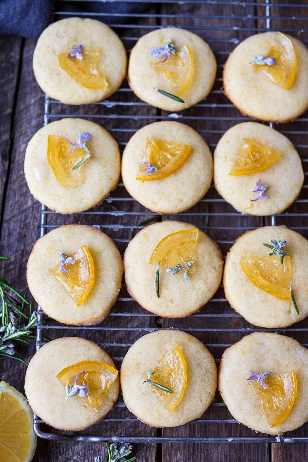 lemon cookies on cooling rack with candied lemons, rosemary, and rosemary flowers, drizzled with lemon glaze.