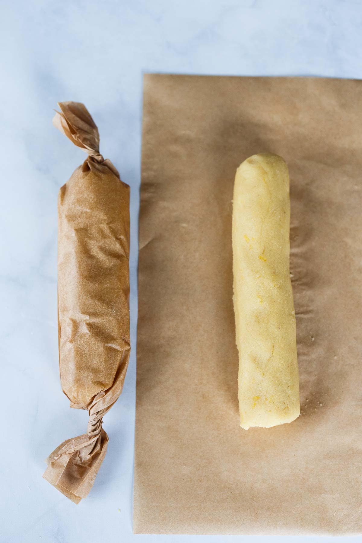 lemon cookie batter rolled into log shape, wrapped up in parchment paper next to roll of lemon cookie batter on top of parchment paper.