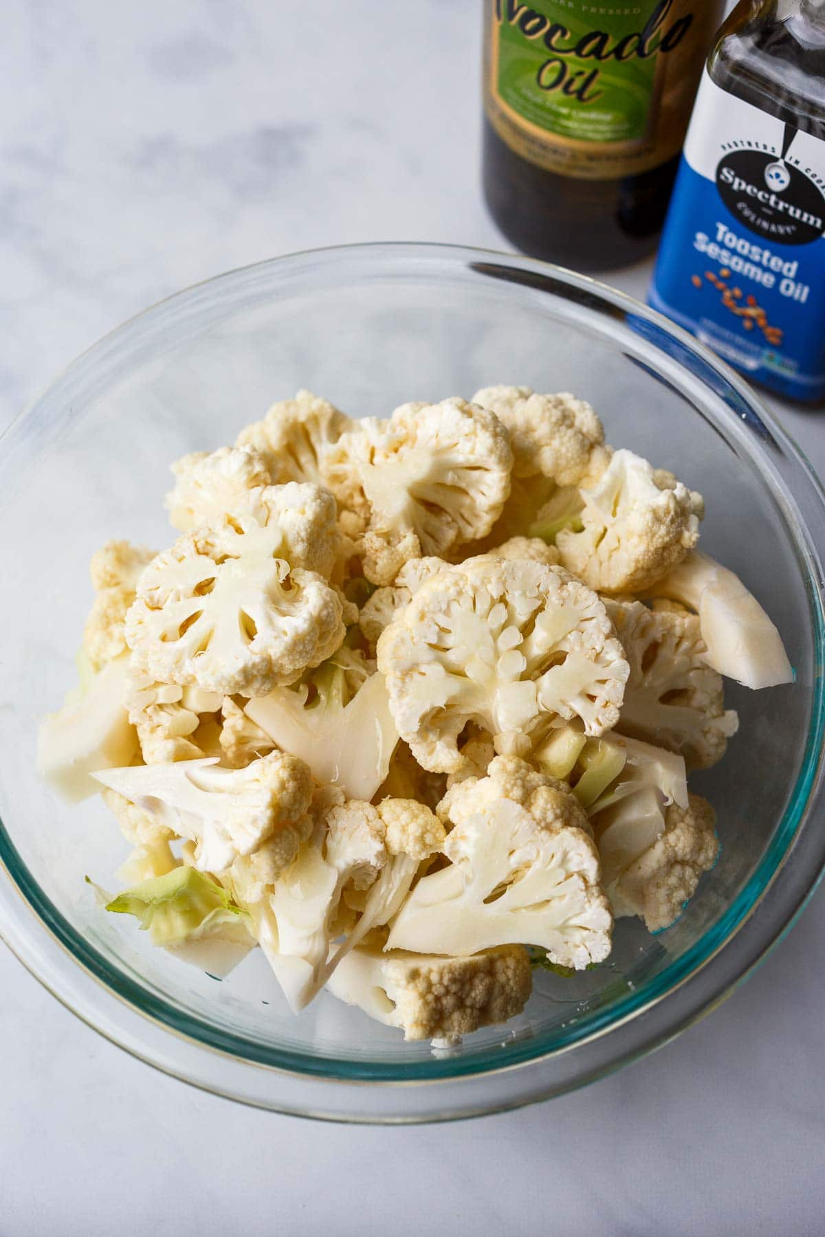 cauliflower florets in glass mixing bowl with toasted sesame oil and olive oil.