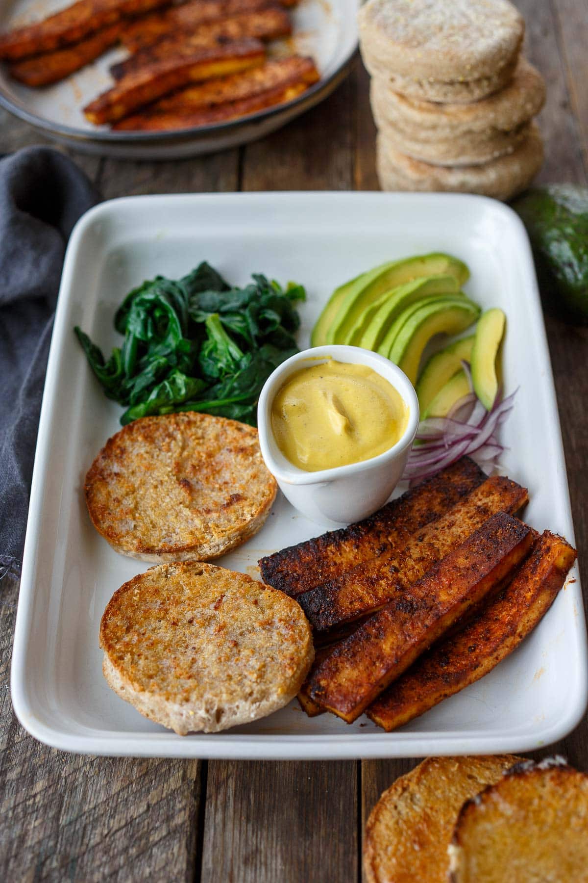 all components for vegan eggs benedict on platter- wilted spinach, avocado, vegan hollandaise sauce, english muffins, tofu bacon.